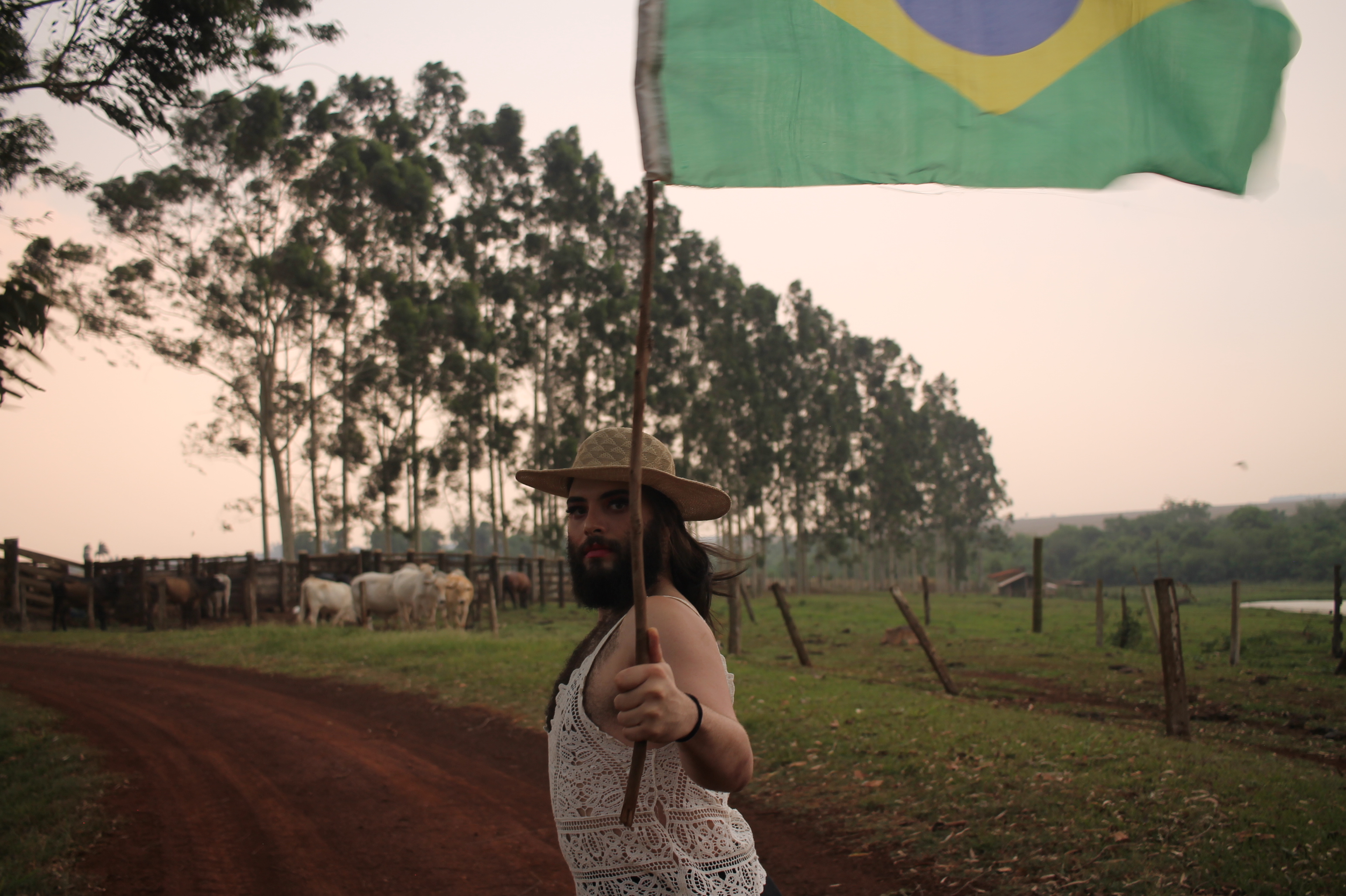  Pessoa com barba, usando chapéu de palha e blusa rendada, segurando uma bandeira do Brasil em uma área rural com árvores e gado ao fundo.